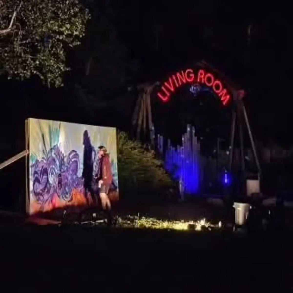 A live-painting panel lit at night beside the illuminated Living Room sign at the Village