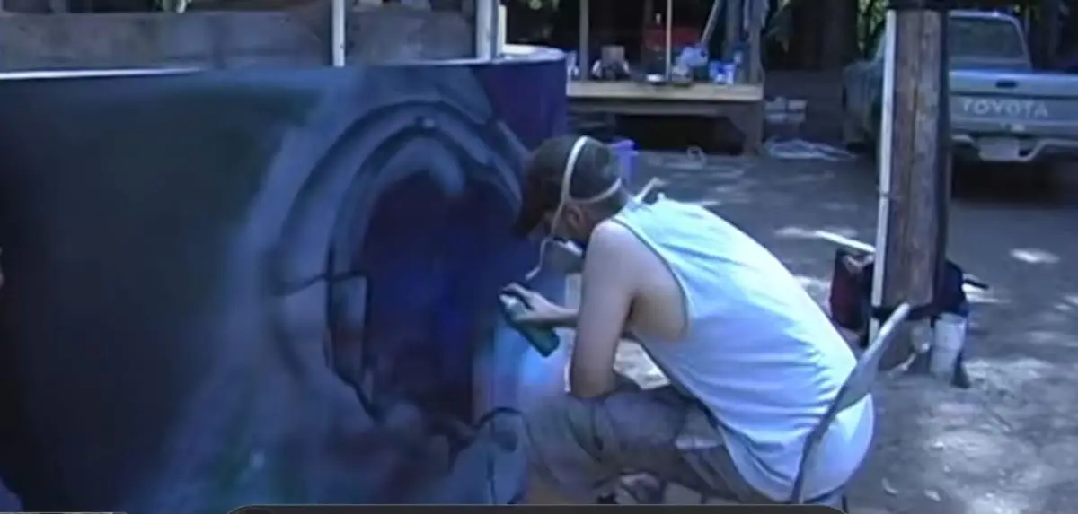 A younger Andreas Tiedemann crouching in a driveway, spraying a blue and purple panel while wearing a respirator